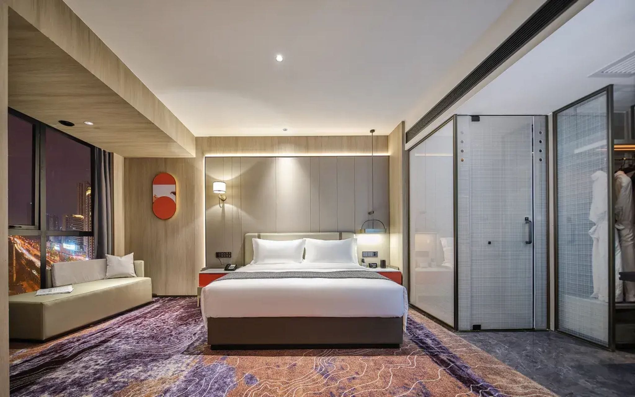 A modern hotel room with a large bed, a seating area, a wardrobe, and a city view.