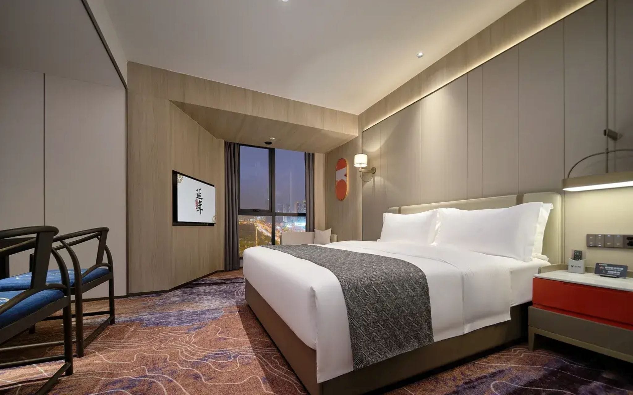 Habitación de hotel con cama, mesita de noche, silla y vistas a la ciudad a través del ventanal.