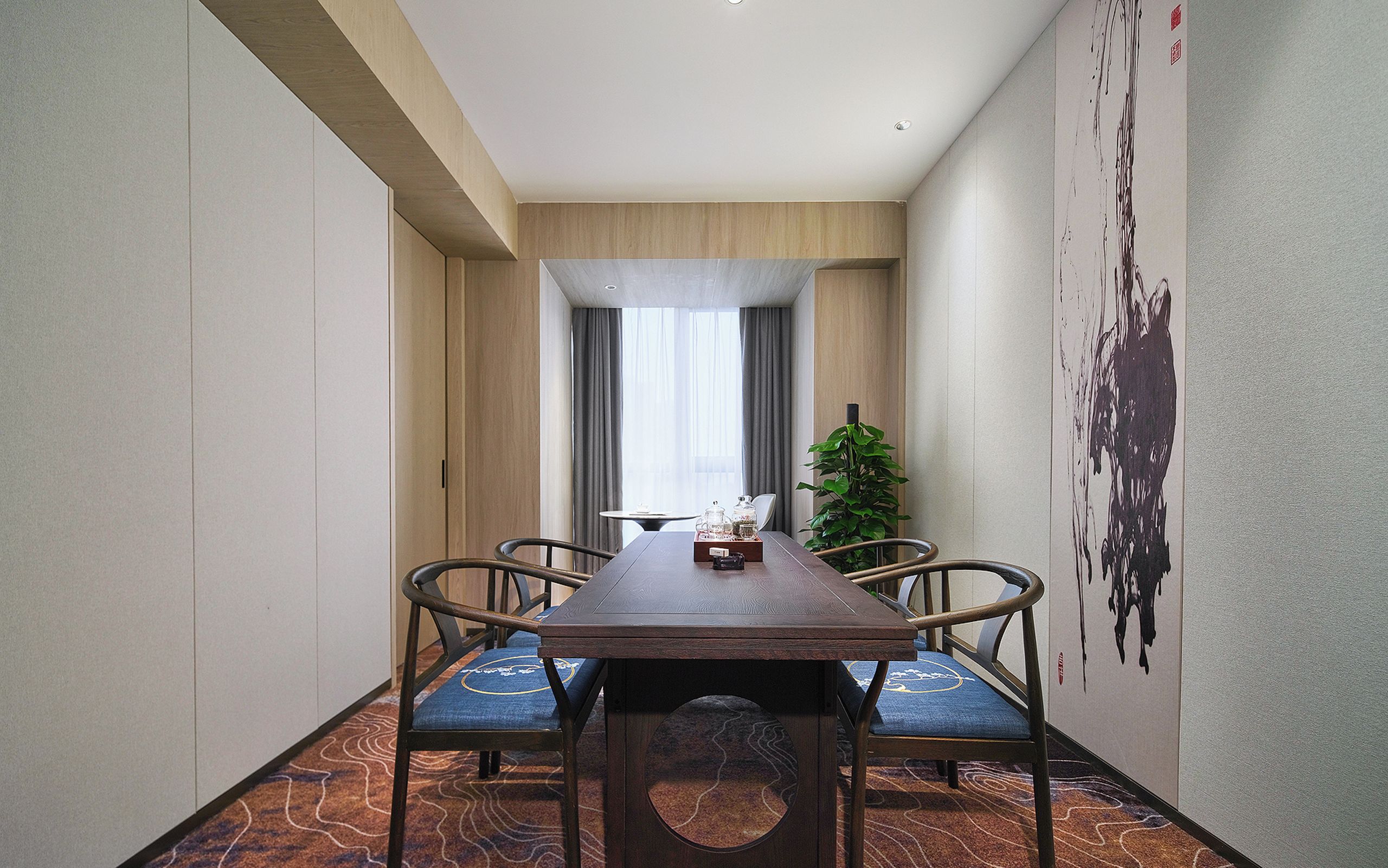 Un comedor de hotel con una larga mesa de madera, cuatro sillas, una planta en maceta y un cuadro abstracto grande en la pared.