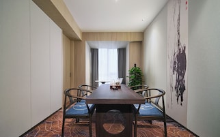Un comedor de hotel con una larga mesa de madera, cuatro sillas, una planta en maceta y un cuadro abstracto grande en la pared.