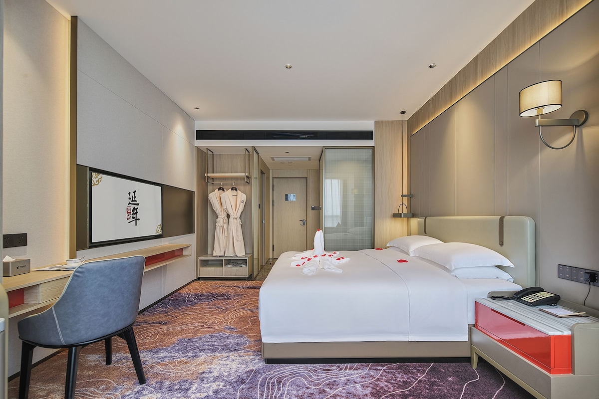 A hotel room with a bed, bedside table, chair, desk, and a bathroom visible through an open door.