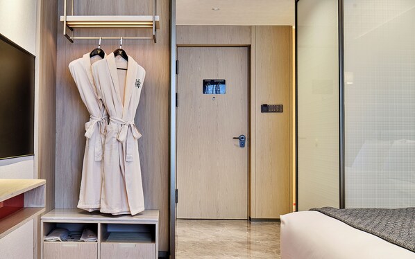A hotel room with a bed, a robe rack with two robes, a door with a keycard slot, and a small shelf with items.