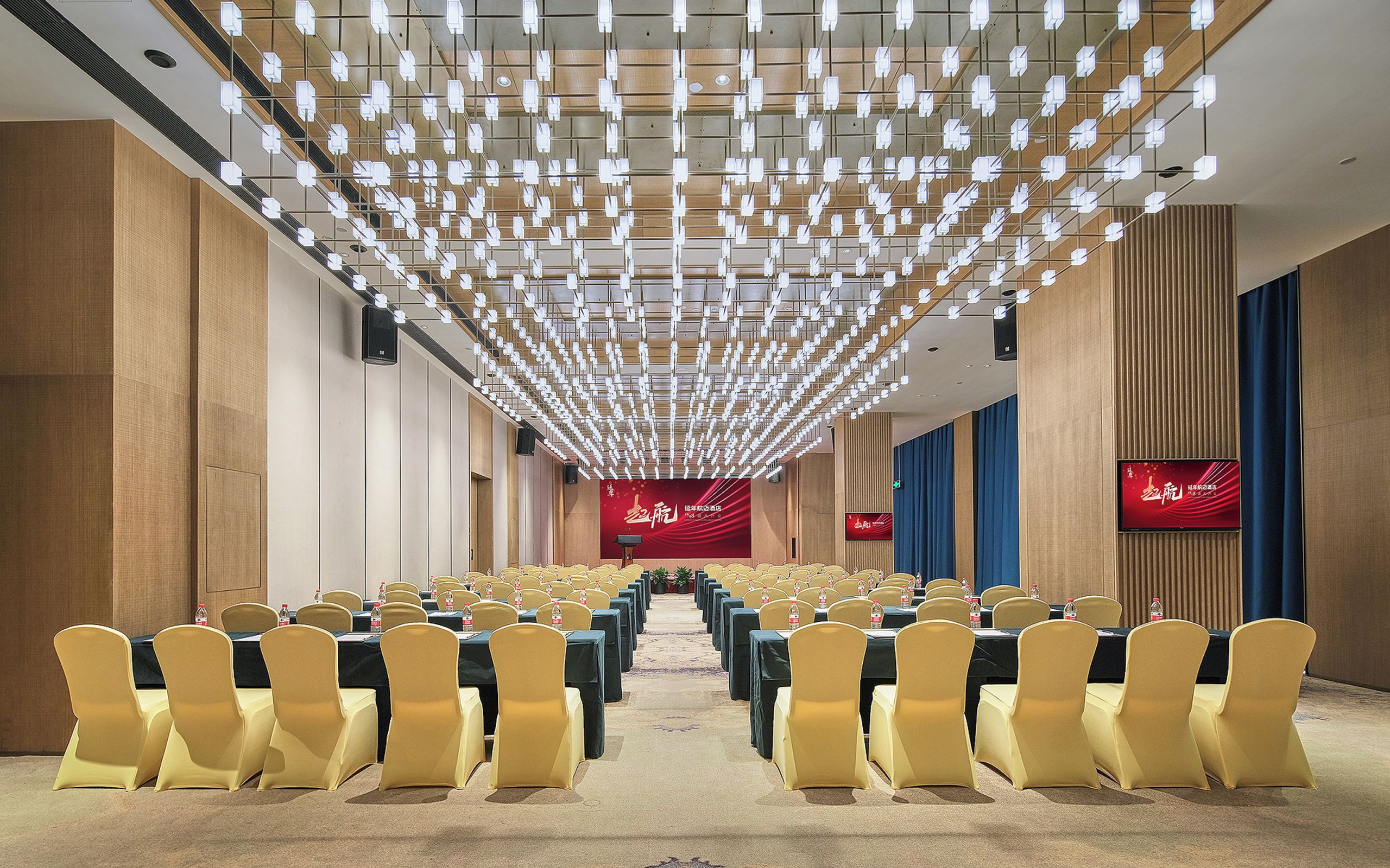 A conference room with rows of tables and chairs, a red curtain backdrop, and a modern ceiling light fixture.
