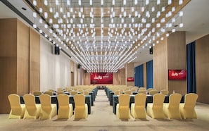 Un salón de conferencias con filas de mesas y sillas, un telón rojo de fondo y una lámpara de techo moderna.