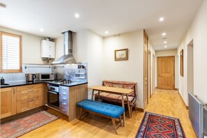 A compact kitchen with a stove, microwave, and cabinets, a dining area with a bench and table, and a hallway leading to a door. - Stylish Notting Hill Two Bed near Bayswater (London)