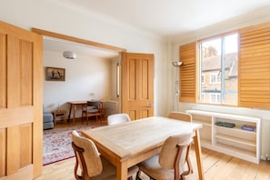 Ein Zimmer mit einem Holztisch, Stühlen, einer Heizung, einem Fenster mit Jalousien und einem Bücherregal.