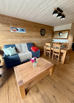 A cozy living room with a wooden ceiling, a blue sofa, a wooden coffee table, and a dining area with wooden chairs and a table. - Chamrousse CAP 2000 Apartment (Chamrousse)