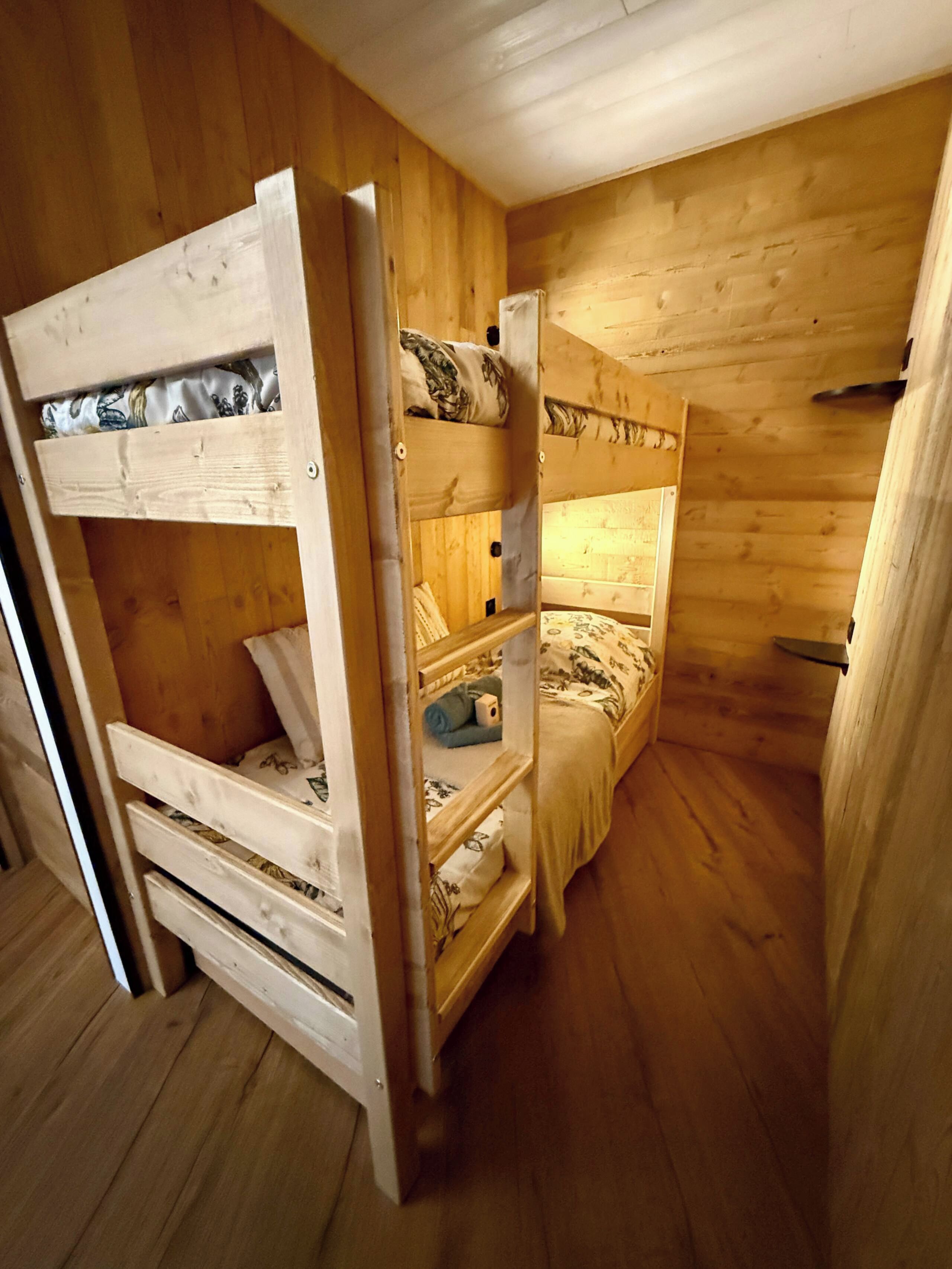 A bunk bed room with wooden walls and floors, a single bed with a patterned bedspread, and a lower bunk with a white headboard.