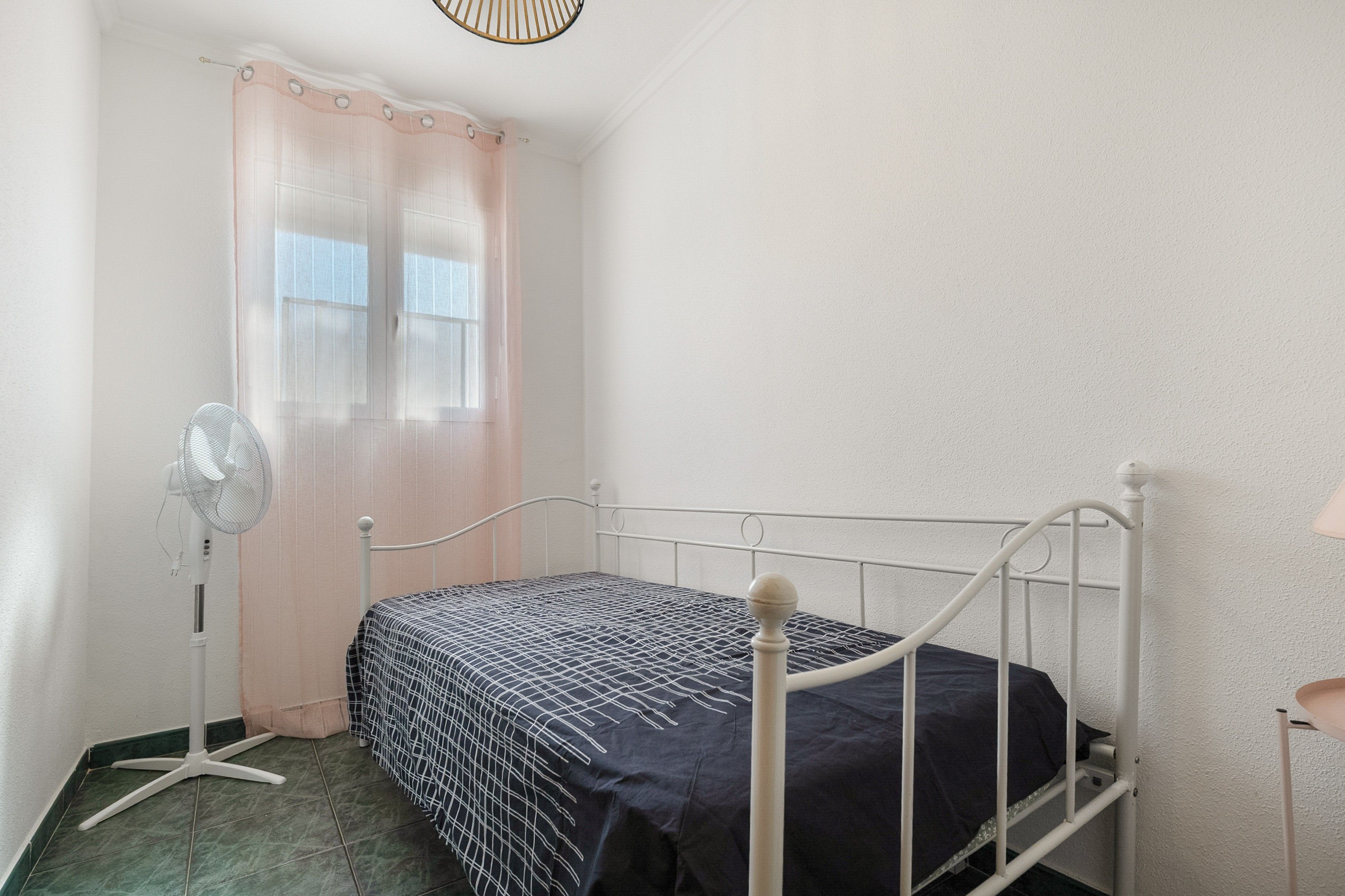 Ein kleines Zimmer mit einem Einzelbett, Metallgestell, einem Standventilator und einem Fenster mit durchsichtigen Vorhängen.