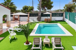 A swimming pool with surrounding lounge chairs and umbrellas, a small table, and a covered area.