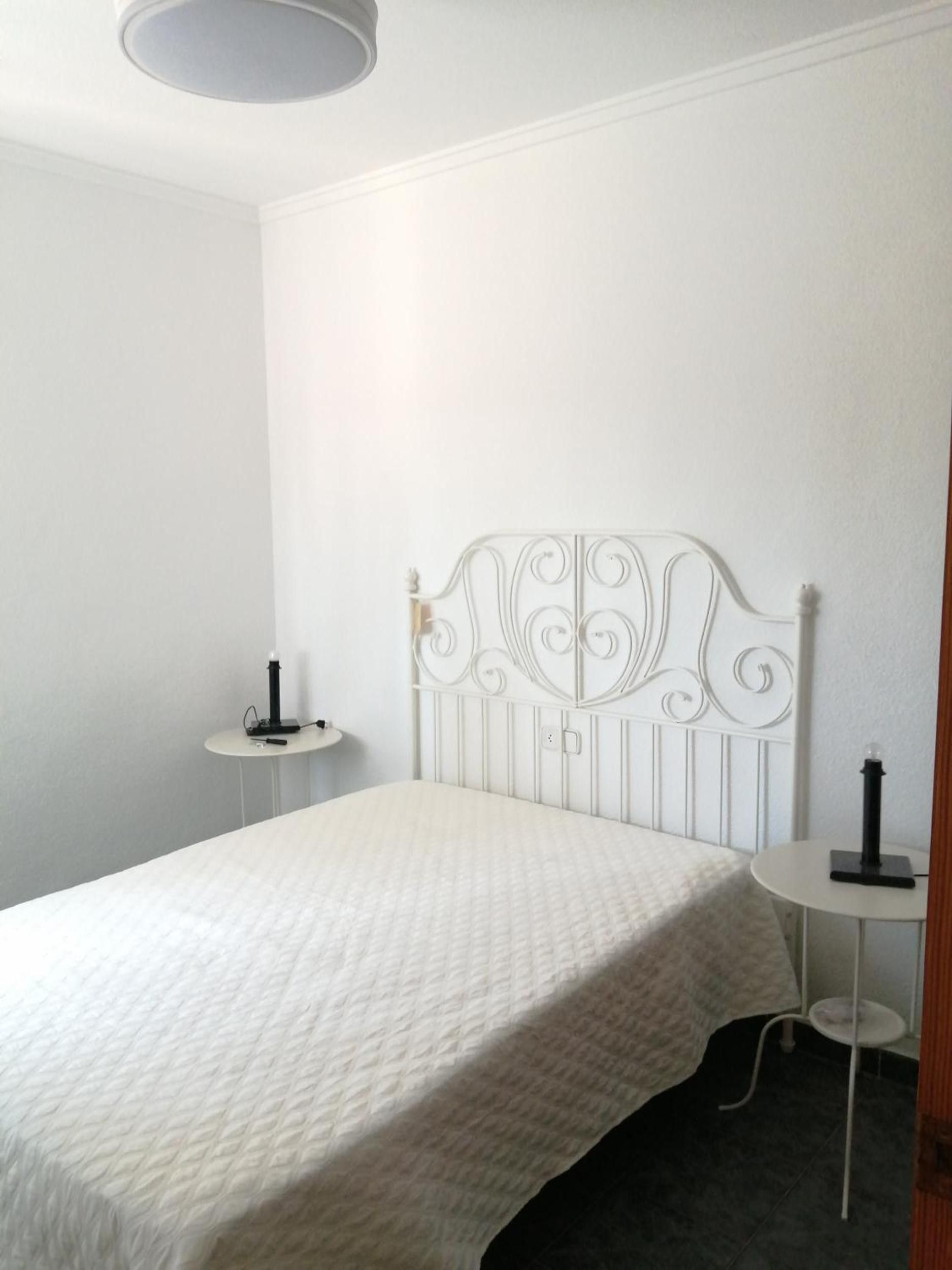 Un dormitorio con cama blanca, cabecera blanca, mesita de noche pequeña y una mesita auxiliar.