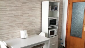 Un angolo cottura con piano di lavoro bianco, un forno a microonde e un frigorifero. C'è un tavolo bianco con due sedie e una panca. La stanza ha un pavimento piastrellato e una porta in legno con un pannello di vetro satinato.