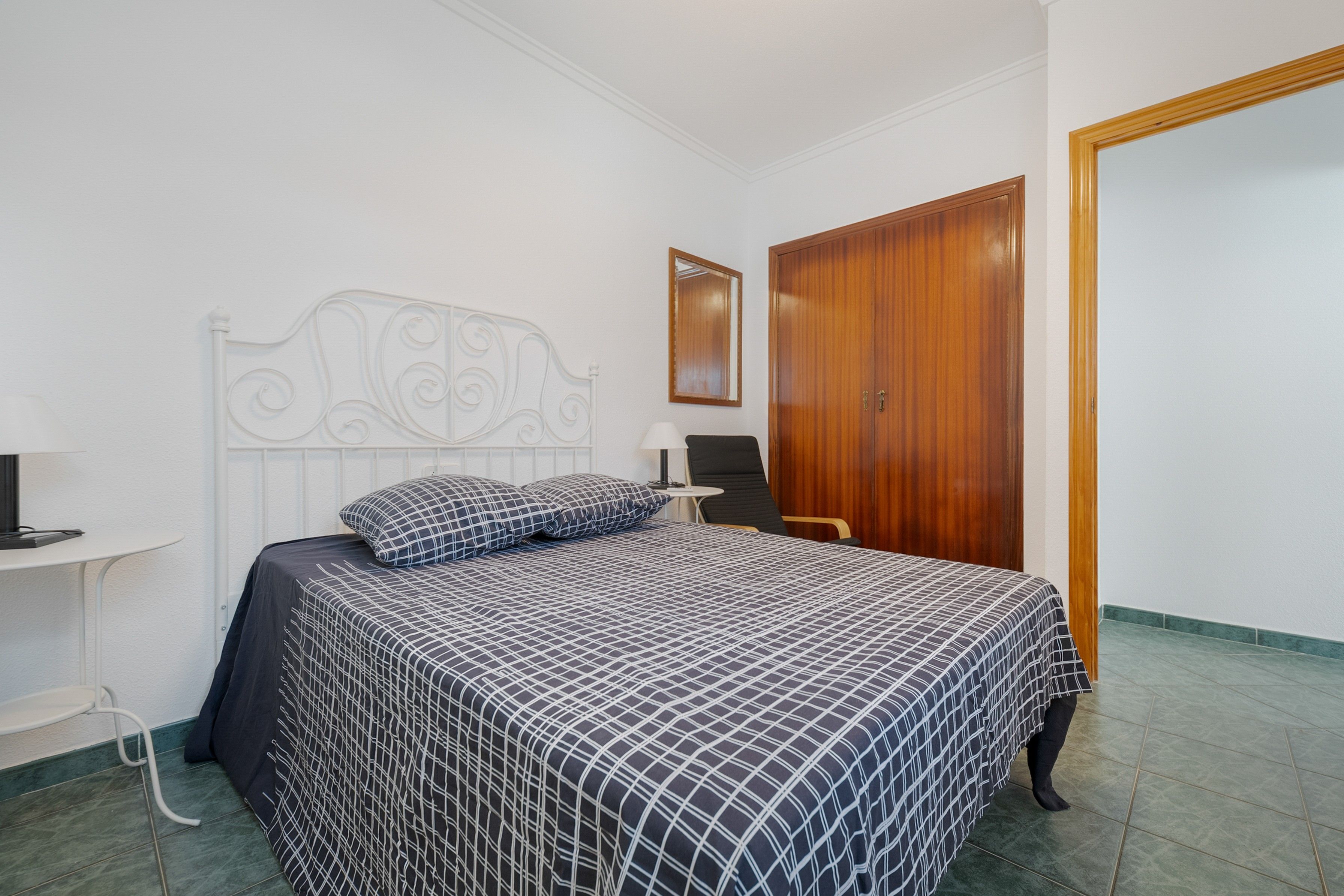 A bedroom with a bed, a wooden wardrobe, a bedside table, and a mirror on the wall.