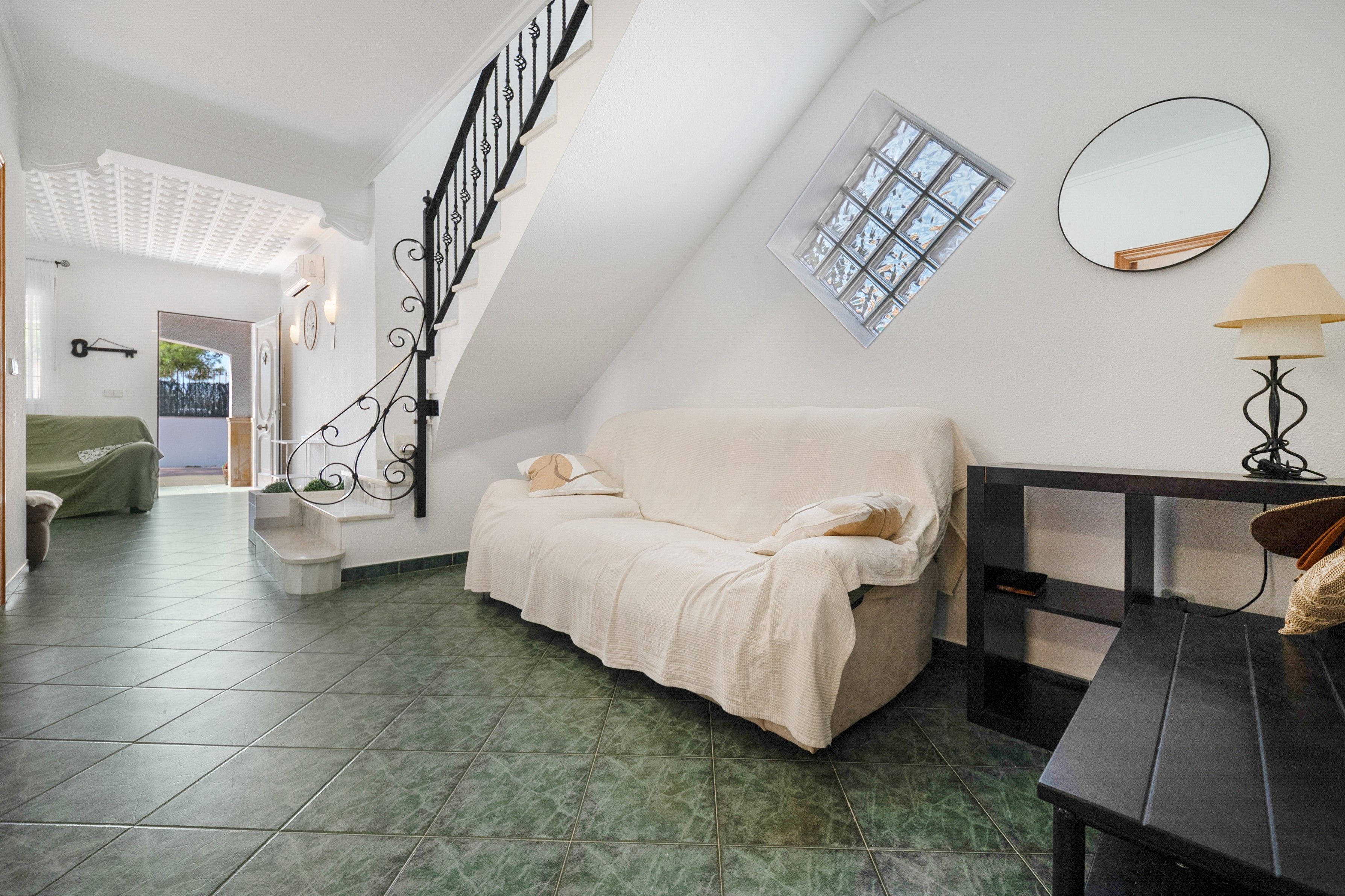 A room with a high ceiling, a staircase with black railings, a sofa with pillows, a small table with a lamp, and a window with a view.