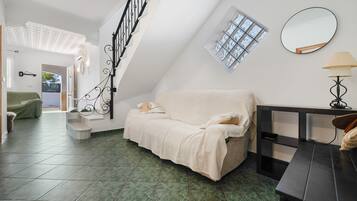 A room with a high ceiling, a staircase with black railings, a sofa with pillows, a small table with a lamp, and a window with a view.