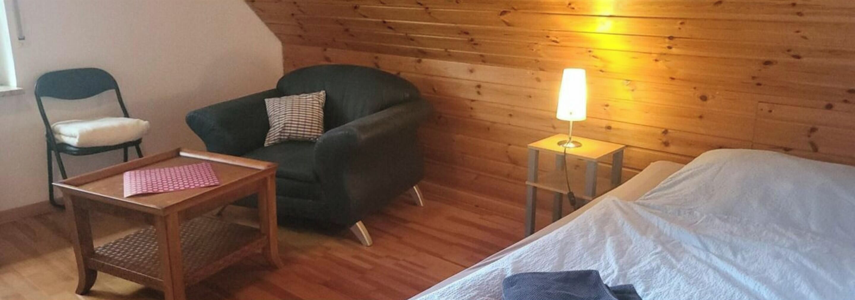 Ein kleines, gemütliches Zimmer mit einem Einzelbett, einem Sessel, einem kleinen Tisch und einer Nachttischlampe.