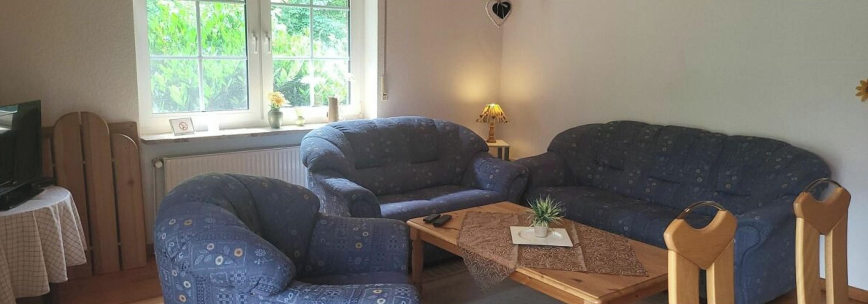 Un salon comprenant un canapé, un fauteuil et une table basse, une télévision et une fenêtre donnant sur un paysage verdoyant.