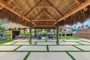 A covered outdoor area with a thatched roof, concrete flooring with grass borders, and a view of a residential area in the background. - Modern Waterfront Retreat Oversize outdoor Tiki, kitchen and heated SPA (Fort Lauderdale)