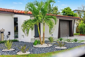 A modern house with a white exterior, a red-tiled roof, and a landscaped garden with palm trees and various plants. - Modern Waterfront Retreat Oversize outdoor Tiki, kitchen and heated SPA (Fort Lauderdale)