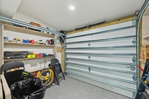 A garage with a large, open door, shelves with tools and supplies, and a workbench with a fan and trash bag. - Modern Waterfront Retreat Oversize outdoor Tiki, kitchen and heated SPA (Fort Lauderdale)