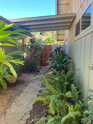 A garden with a stone pathway leading to a wooden gate, surrounded by various plants and a covered area. - Hale Manu i ke Kai (Kihei)