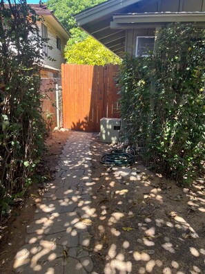 A paved pathway leading to a wooden gate, surrounded by dense foliage and a utility box. - Hale Manu i ke Kai (Kihei)