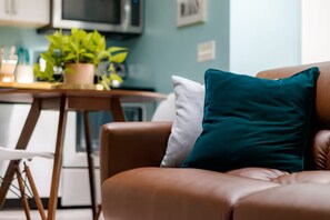 A brown leather sofa with teal and white cushions, a wooden chair, a small table with a plant, and a kitchen area in the background. - Private Guesthouse Near Beach & Downtown (St. Petersburg)