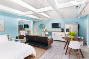 Ein modernes Schlafzimmer mit Bett, einer Couch, einem Esstisch mit Stühlen, einer Küchenzeile mit Kühlschrank und einem an der Wand befestigten Fernseher.