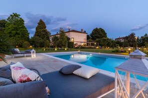 Ein Swimmingpool mit Liegestühlen und Kissen, umgeben von einem gepflegten Rasen und Bäumen.