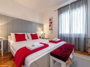 A hotel room with a large bed, a bedside table, a lamp, and a view of the outdoors through sheer curtains. - Three Bedroom Apartment with Balcony (Rogoznica)
