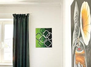 Une pièce avec une fenêtre, un tableau abstrait aux tons verts et une vue partielle sur une planche de surf.