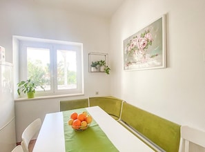 Un espace repas comprenant une table, des chaises, une fenêtre, un banc vert avec coussin, une plante fixée au mur et un tableau encadré.