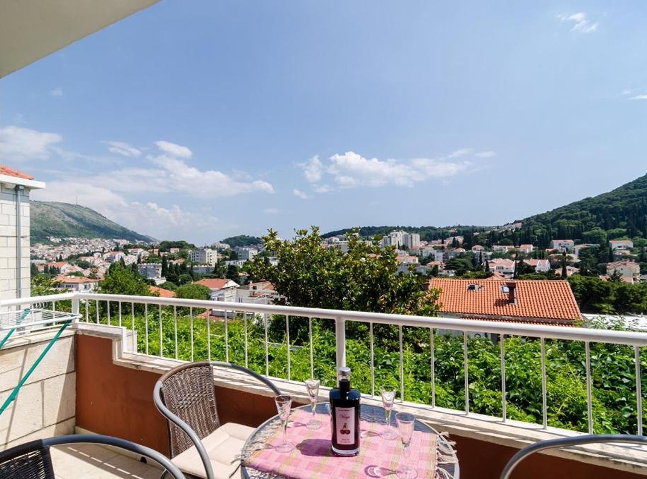 A balcony with a table, chairs, and a bottle of wine, overlooking a town with buildings and hills.