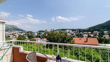 A balcony with a table, chairs, and a bottle of wine, overlooking a town with buildings and hills.