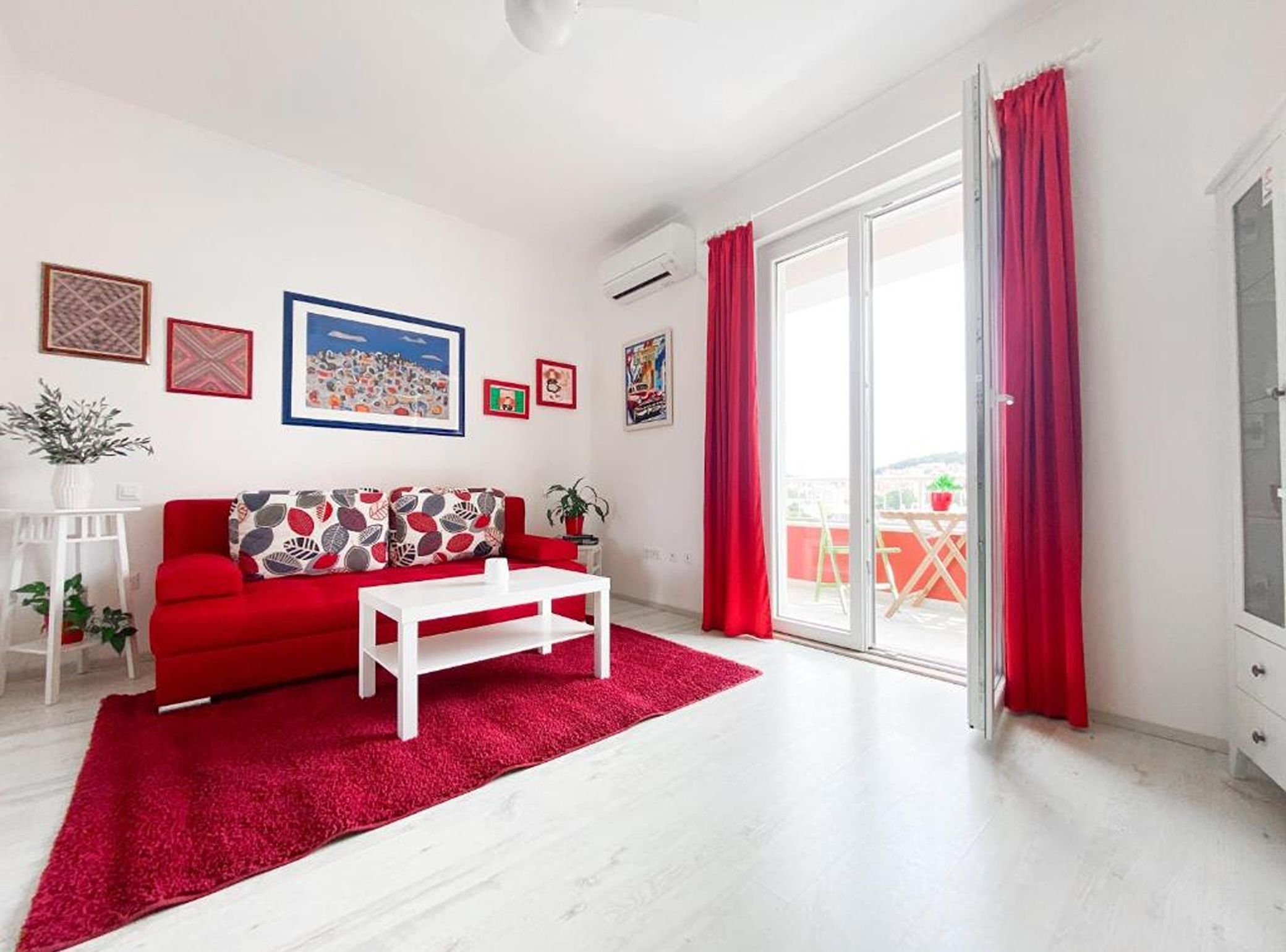 A living room with a red sofa, white coffee table, and a red rug. There are framed pictures on the wall, a potted plant, and a sliding glass door leading to an outdoor area.