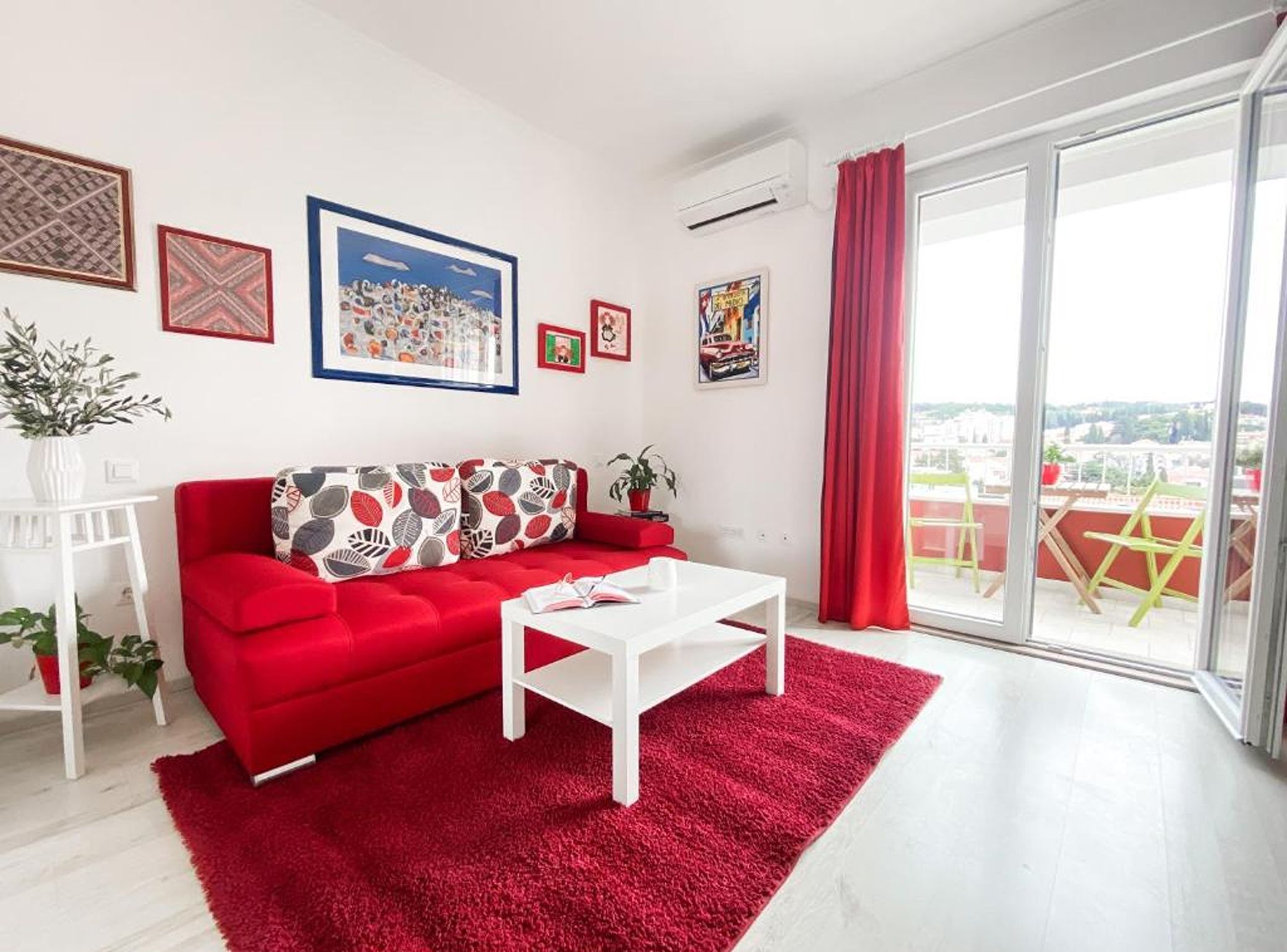 A modern living room with a red sofa, white coffee table, and a red rug. There are framed pictures on the wall, a potted plant, and a view through the large window.