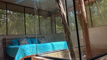 A modern bedroom with a large window, a bed with blue bedding, and a tree trunk integrated into the room's design.