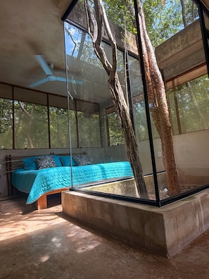A modern bedroom with a large window, a bed with blue bedding, and a tree trunk integrated into the room's design. - Hotel El Encanto Chichen Itzá (Sudzal)