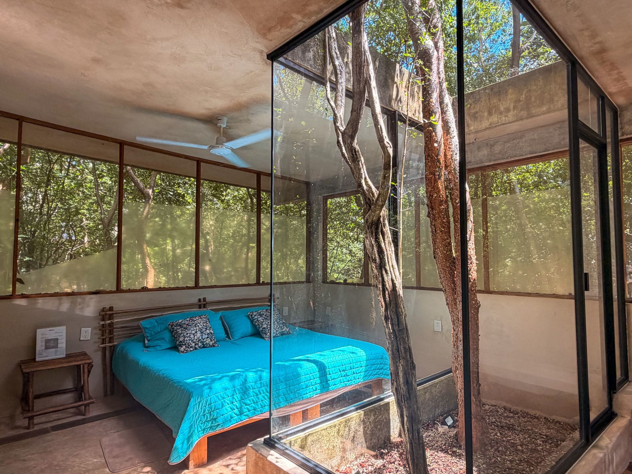 Een slaapkamer met een bed, een houten nachtkastje en een groot raam met uitzicht op bomen.
