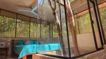 A modern bedroom with a large window, a bed with blue bedding, and a tree trunk in the center.