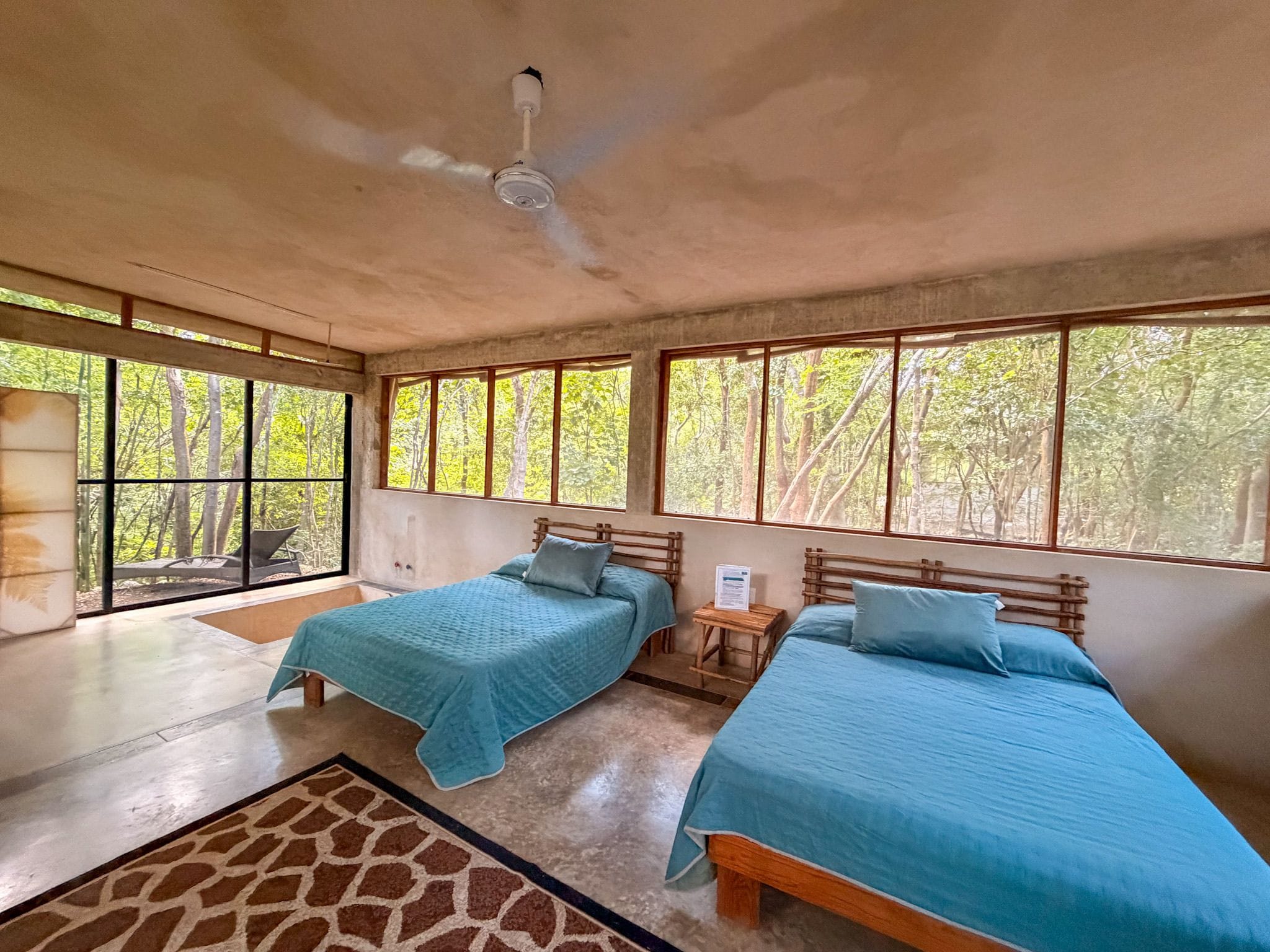 A room with two beds, a ceiling fan, and large windows with a forest view.