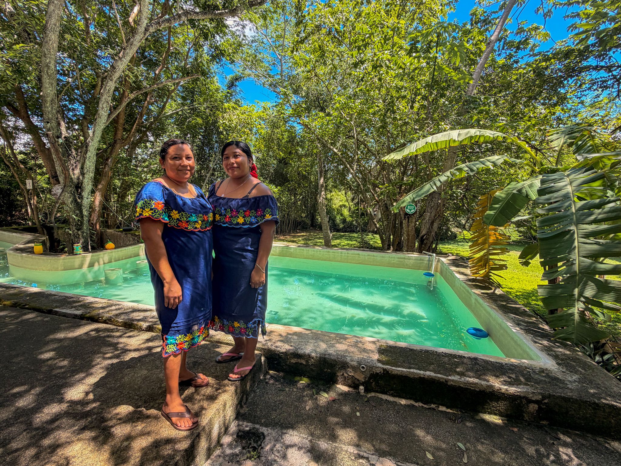 Dos personas junto a una piscina con un jardín tropical al fondo.