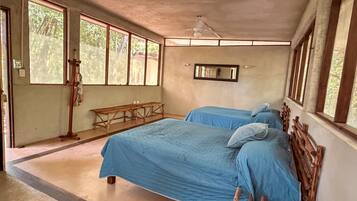 A room with two beds, a wooden bench, a ceiling fan, and large windows.