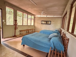 Una habitació amb dos llits, una banca de fusta, un ventilador de sostre i grans finestres.