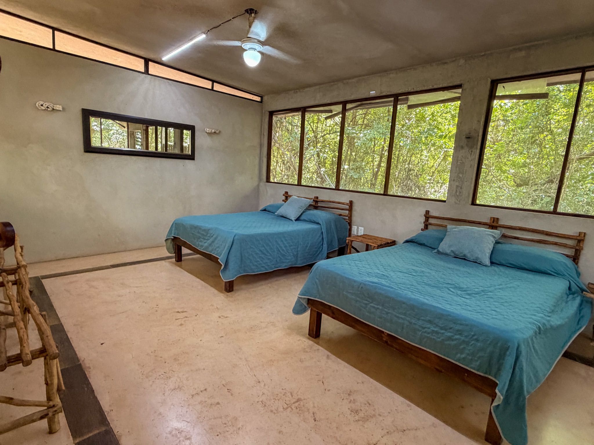 Quarto com duas camas de madeira, um ventilador de teto e janelas com vista para árvores.