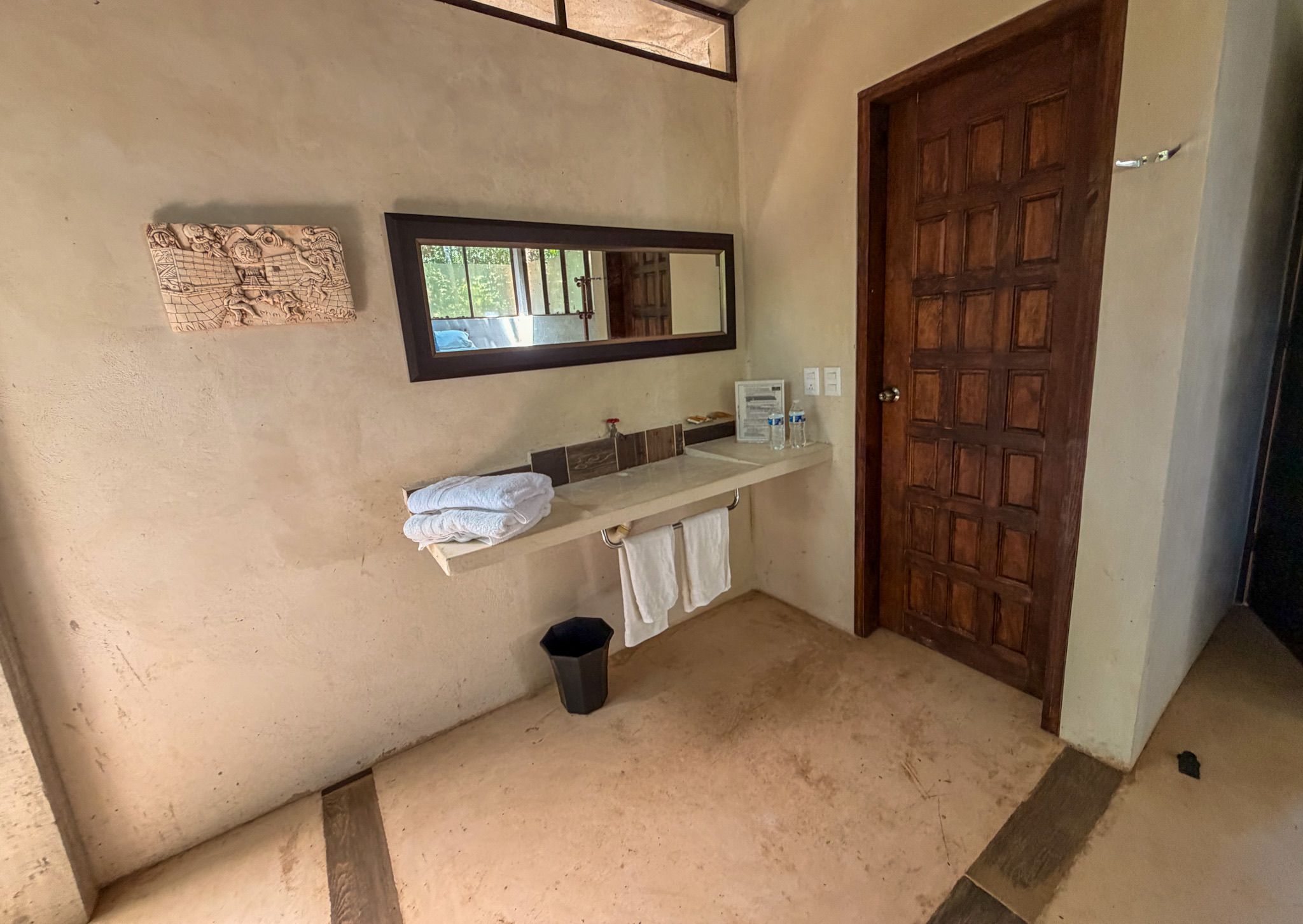 Un baño con puerta de madera, un espejo, toallero y un cubo de basura.