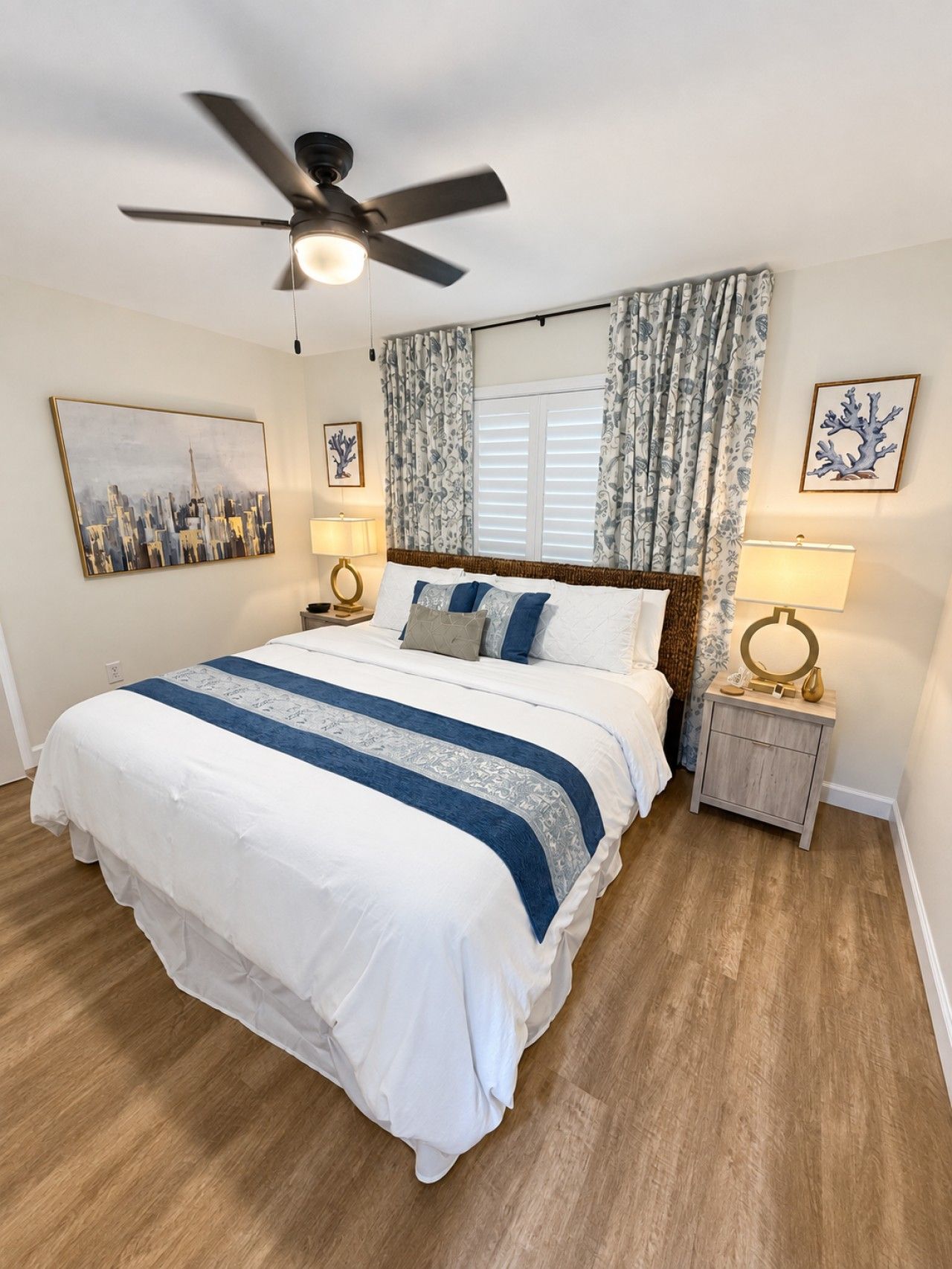 A bedroom with a bed, a ceiling fan, a nightstand with a lamp, and a window with curtains.