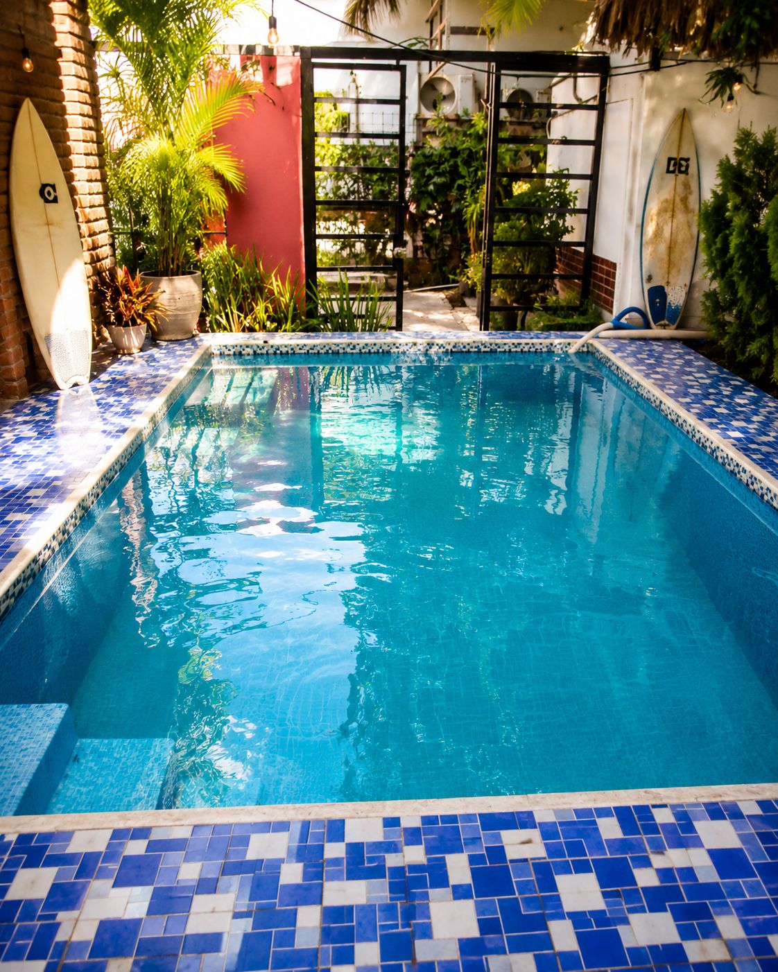 Una piscina rectangular con mosaicos azules y blancos, rodeada de follaje y una pared roja.