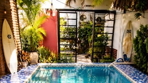 Una piscina rectangular con mosaicos azules y blancos, rodeada de follaje y una pared roja.