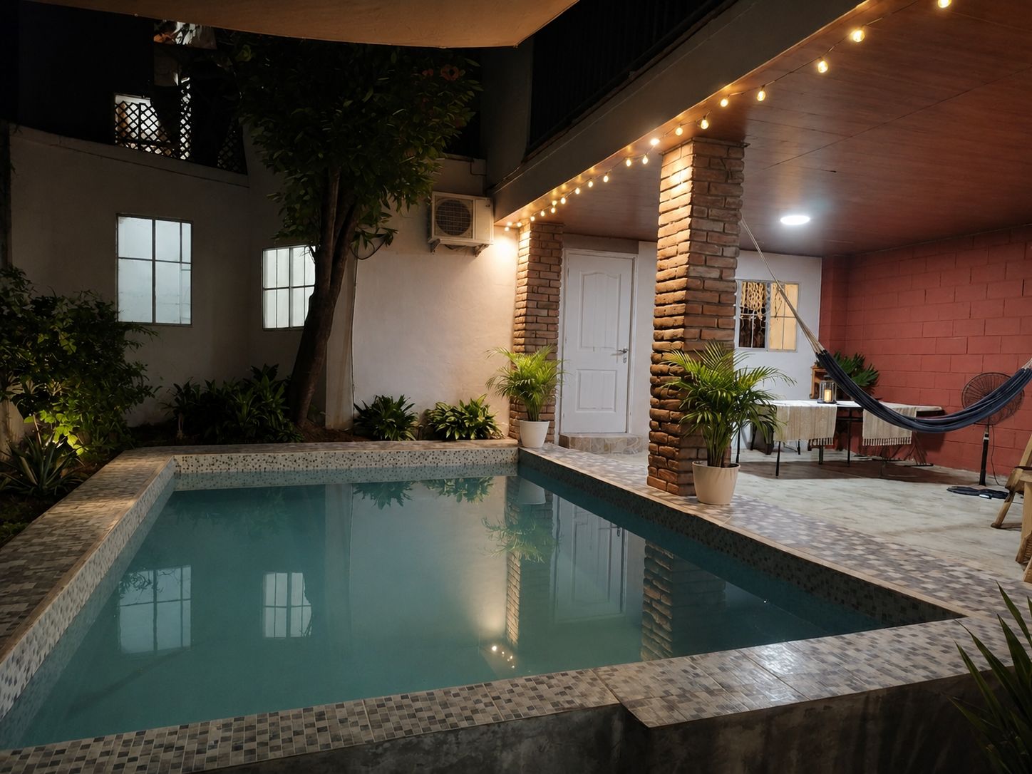 Una piscina con borde de azulejos, rodeada de plantas en macetas y una hamaca.